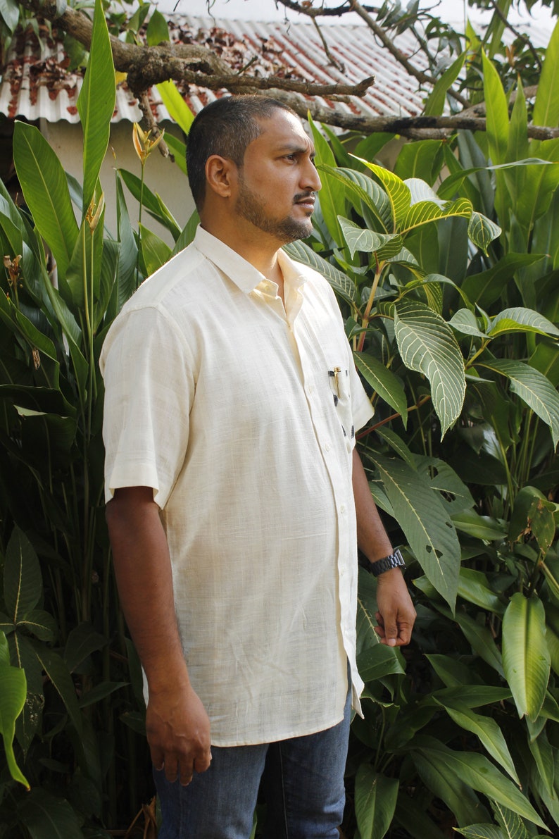 May include: A light beige short-sleeved button-down shirt with a pocket, paired with blue jeans. The shirt is worn by a person standing in front of lush green foliage. A watch is visible on the person's wrist.