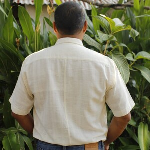 May include: A person wearing a cream-colored short-sleeved shirt and blue jeans. The shirt has a button-down collar and a textured appearance. The jeans have a brown leather patch on the back pocket.