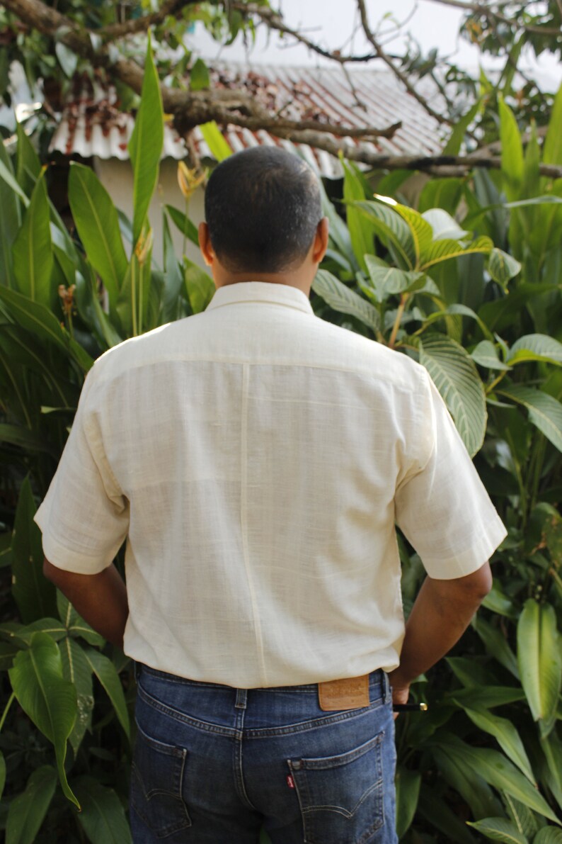 May include: A person wearing a cream-colored short-sleeved shirt and blue jeans. The shirt has a button-down collar and a textured appearance. The jeans have a brown leather patch on the back pocket.