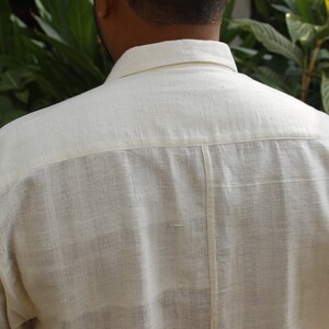 May include: Back view of a cream-colored button-down shirt. The shirt has a classic collar and short sleeves. The fabric appears to be a textured linen or cotton blend. The shirt is a neutral color, suitable for various outfits.