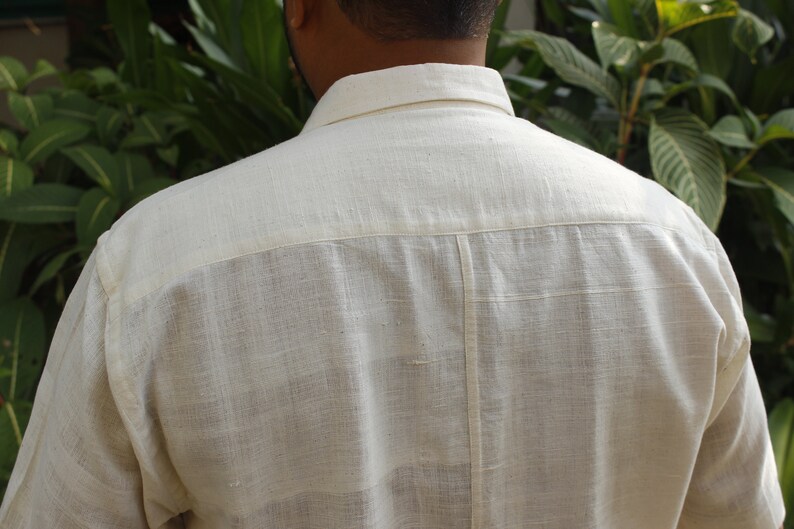 May include: Back view of a cream-colored button-down shirt. The shirt has a classic collar and short sleeves. The fabric appears to be a textured linen or cotton blend. The shirt is a neutral color, suitable for various outfits.