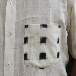 May include: Off-white short-sleeved shirt with a pocket featuring a black and white geometric pattern. The shirt has a button-down front and a classic collar. The fabric appears to be a textured, lightweight material.