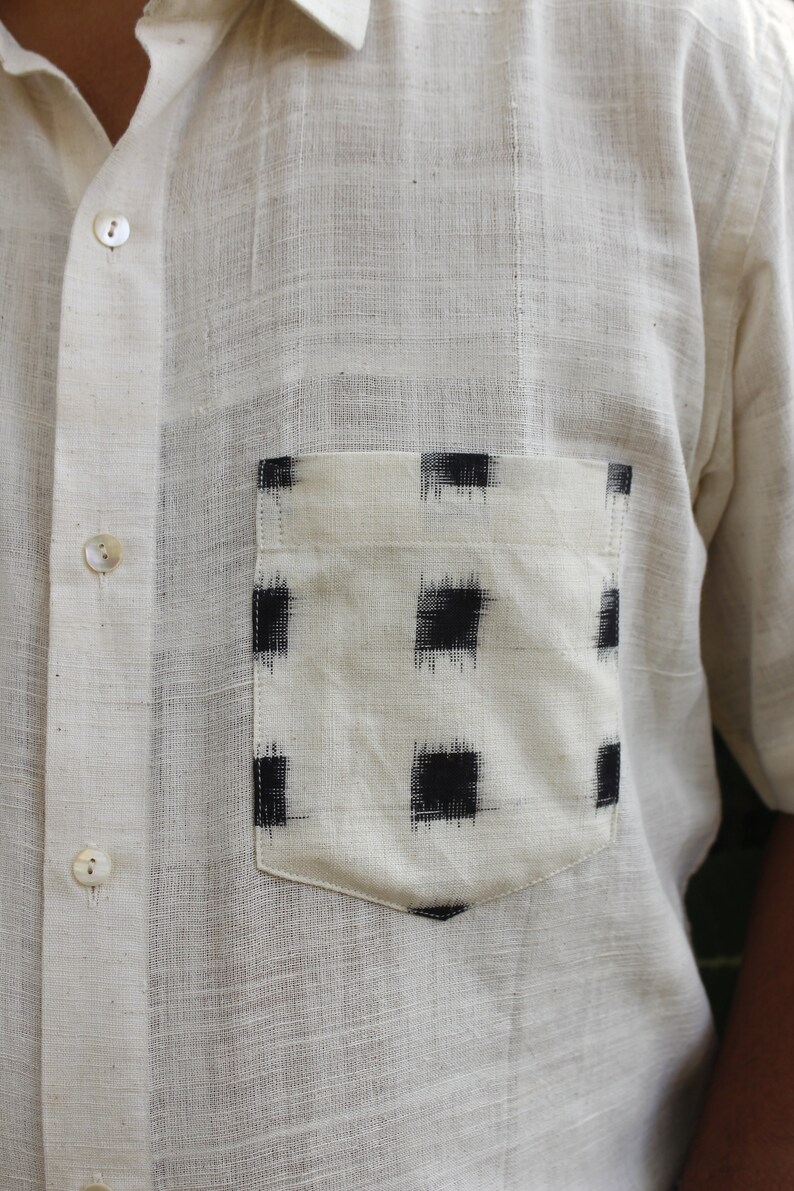 May include: Off-white short-sleeved shirt with a pocket featuring a black and white geometric pattern. The shirt has a button-down front and a classic collar. The fabric appears to be a textured, lightweight material.