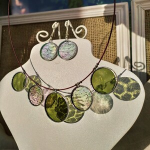 May include: A necklace and earrings set with round, iridescent glass pendants. The necklace has five pendants in shades of green and white, while the earrings have two pendants in shades of pink and white.