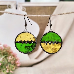 May include: A pair of earrings with round, green and yellow glass pieces. The glass pieces are separated by a thin, black metal band with a wavy design.