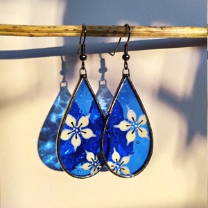 Könnte beinhalten: Ein Paar tropfenförmige Ohrringe mit blauem Hintergrund und weißen Blumenmustern. Die Ohrringe sind aus Glas und haben einen silberfarbenen Metallhaken.