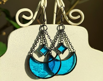 May include: A pair of blue crescent moon earrings with a diamond-shaped accent and a chain dangle. The earrings are made of stained glass and have a black metal frame.