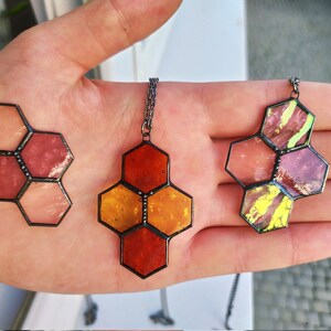 May include: Three stained glass hexagon pendants in shades of red, orange, and pink. Each pendant is connected to a silver chain. The glass has a textured appearance, and the metal frames are dark.