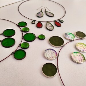 May include: A set of three necklaces and earrings featuring round and teardrop-shaped glass pendants in green, red, and white with iridescent patterns. The necklaces are made with thin, brown cords.