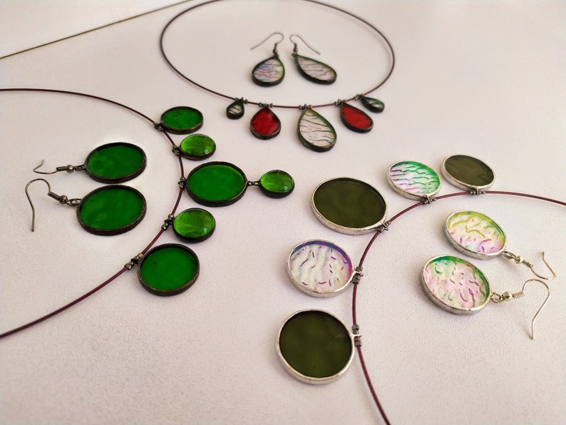 May include: A set of three necklaces and earrings featuring round and teardrop-shaped glass pendants in green, red, and white with iridescent patterns. The necklaces are made with thin, brown cords.
