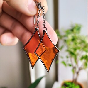 Op de afbeelding: Een paar oranje gebrandschilderde oorbellen met een diamantvorm en een zilveren ketting.