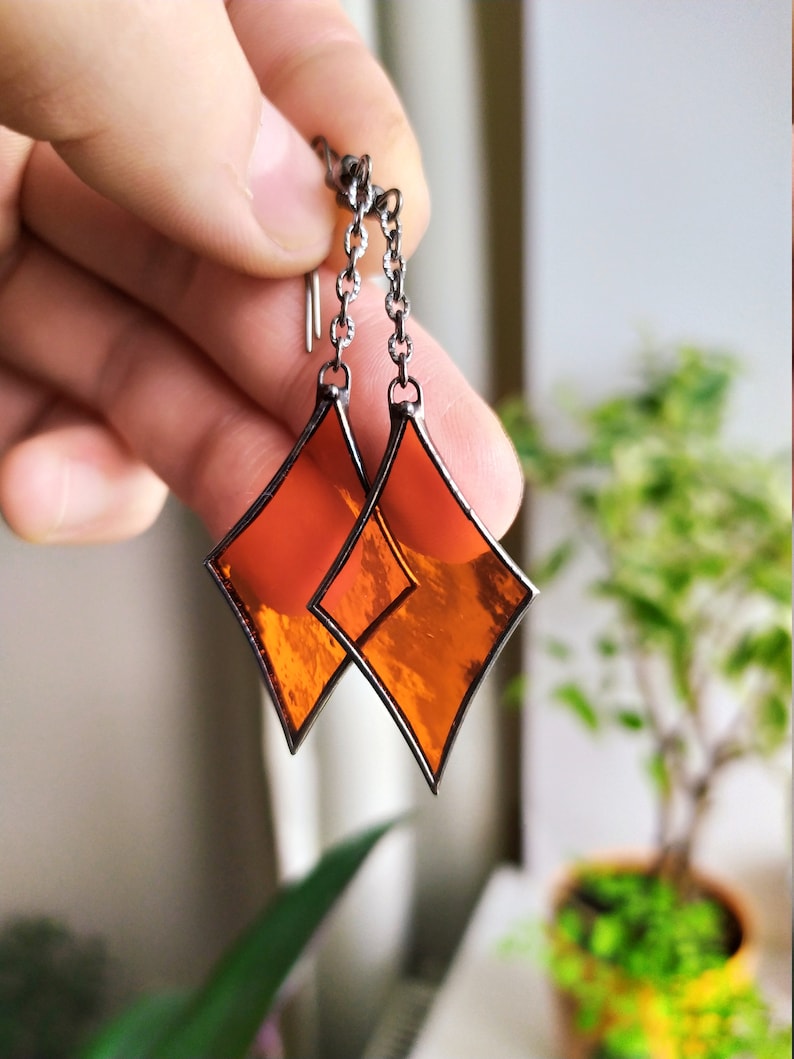 Op de afbeelding: Een paar oranje gebrandschilderde oorbellen met een diamantvorm en een zilveren ketting.