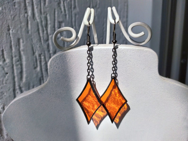 Op de afbeelding: Een paar oranje diamantvormige glazen oorbellen met een zwart metalen frame en ketting. De oorbellen hangen aan een witte displaystandaard.