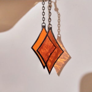 Op de afbeelding: Twee oranje glazen diamantvormen die aan een zilveren ketting hangen. De diamanten zijn op elkaar gelegd.