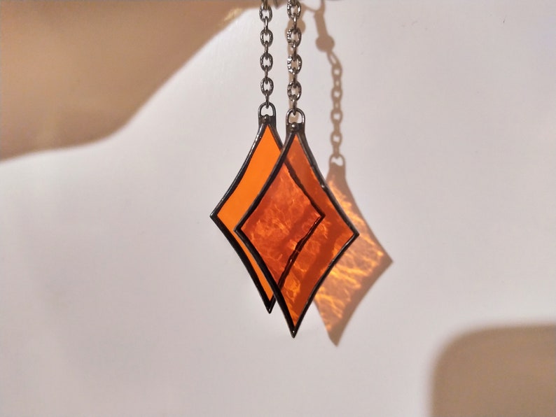 Op de afbeelding: Twee oranje glazen diamantvormen die aan een zilveren ketting hangen. De diamanten zijn op elkaar gelegd.