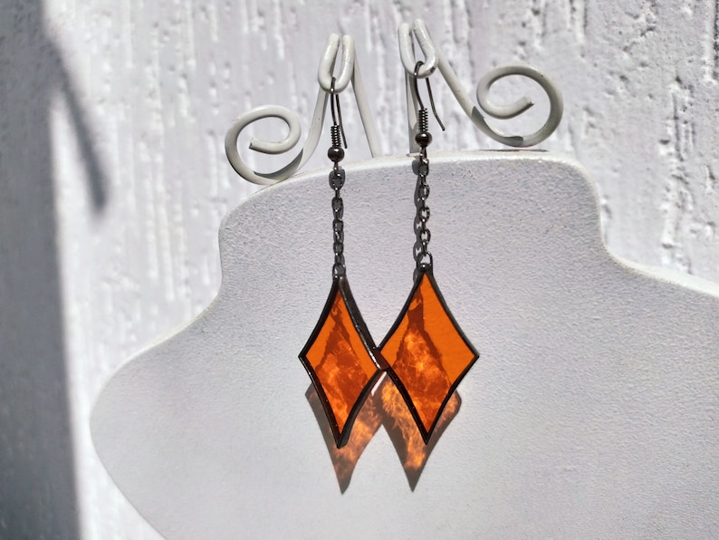 Op de afbeelding: Een paar oranje ruitvormige glas-in-lood oorbellen met zwarte contouren die aan zilveren kettingen hangen.