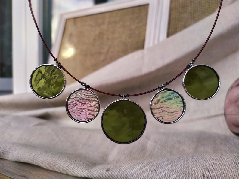 May include: A necklace with five round glass pendants. The pendants are in shades of green, pink, and white. The necklace is made of a thin red cord.