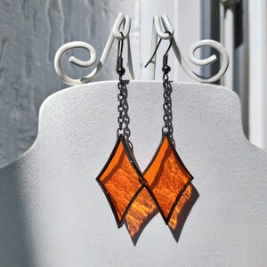 Op de afbeelding: Een paar oranje glazen diamantvormige oorbellen met zwarte metalen frames en kettingen.