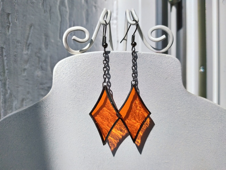 Op de afbeelding: Een paar oranje glazen diamantvormige oorbellen met zwarte metalen frames en kettingen.