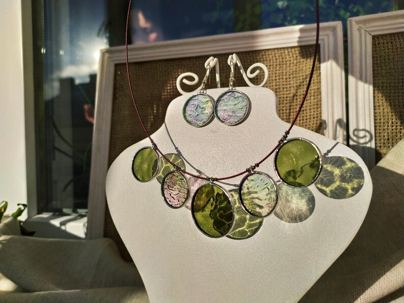 May include: A necklace and earrings set with round, iridescent glass pendants. The necklace has five pendants in shades of green and white, while the earrings have two pendants in shades of pink and white.