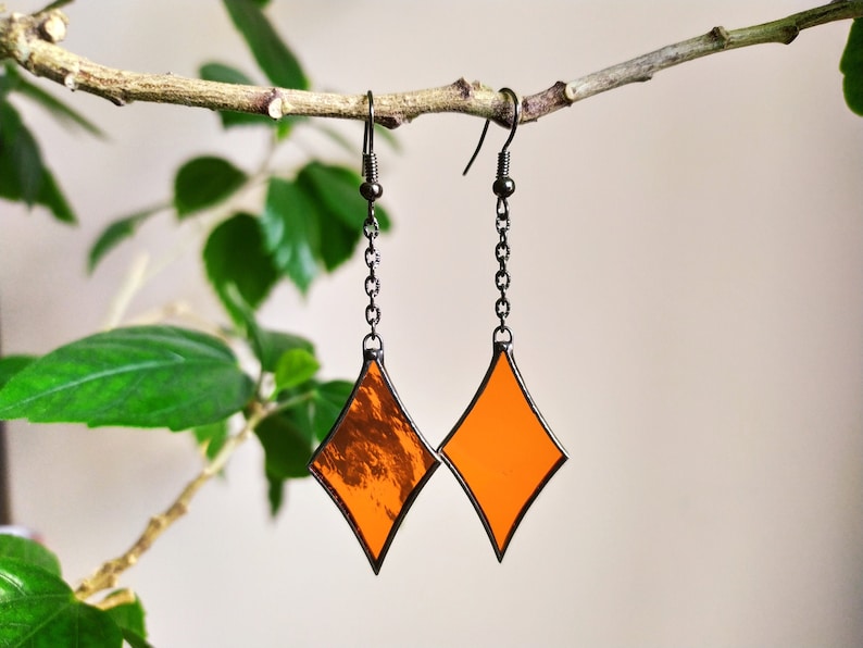 Op de afbeelding: Een paar oranje ruitvormige glas-in-lood oorbellen met zwarte metalen kettingen.