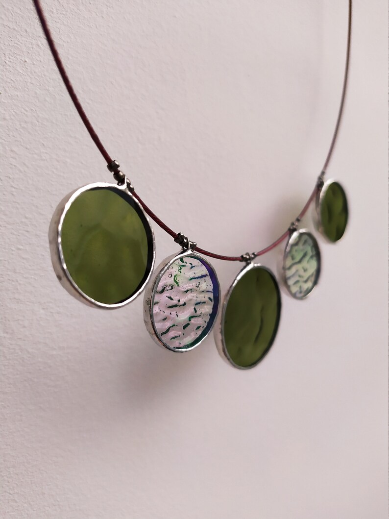 May include: A necklace with five round glass pendants. The pendants are in shades of green and white with a silver trim. The necklace is made of a thin red cord.