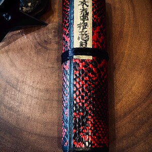 Könnte beinhalten: Zylindrisches Objekt mit einem roten und schwarzen strukturierten Muster. Es hat ein schwarzes Band und einen Holzsockel. Japanischer Text ist auf einem rechteckigen Etikett sichtbar. Ca. 20 cm lang.