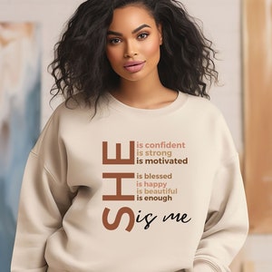 Op de afbeelding: Een beige sweatshirt met de tekst "SHE is confident is strong is motivated is blessed is happy is beautiful is enough is me" gedrukt op de voorkant.