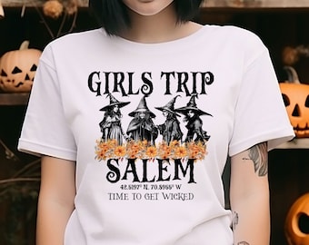 May include: White t-shirt with the text "GIRLS TRIP SALEM" and an illustration of four witches. The shirt also includes the coordinates 42.5197° N, 70.5959° W and the phrase "TIME TO GET WICKED". Pumpkins are in the background.