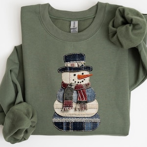 May include: An olive green sweatshirt featuring a snowman design. The snowman has a black hat, a red and blue scarf, and a carrot nose. The snowman is made of layered fabric with a denim-like pattern.