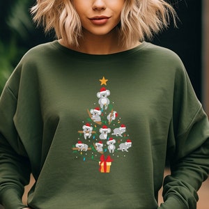 May include: A green sweatshirt with a Christmas tree design featuring koalas wearing Santa hats. The tree is decorated with lights and a red gift box at the base.