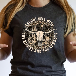 May include: A dark gray t-shirt with a graphic print of a cow skull with a leopard print background. The text "Raisin' Hell With The Hippies And The Cowboys" is printed around the skull.