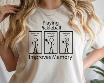 Op de afbeelding: Een lichtbeige t-shirt met de tekst "Playing Pickleball Improves Memory". Het ontwerp toont stokfiguren en tekstballonnen met vragen over het spel. Een humoristisch ontwerp voor pickleball-liefhebbers.