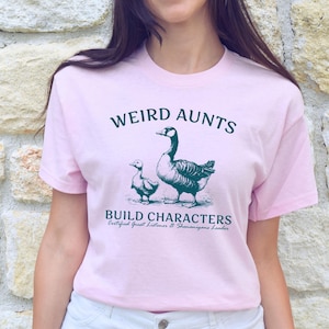 May include: A light pink t-shirt with the text "WEIRD AUNTS BUILD CHARACTERS" in dark green, along with an illustration of a goose and a gosling. The shirt also includes the text "Certified Great Listeners & Shenanigans Leader."