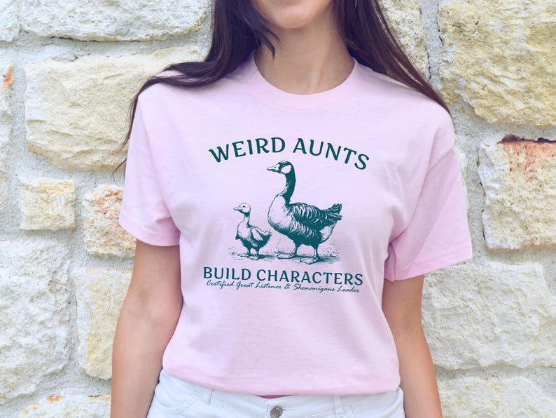 May include: A light pink t-shirt with the text "WEIRD AUNTS BUILD CHARACTERS" in dark green, along with an illustration of a goose and a gosling. The shirt also includes the text "Certified Great Listeners & Shenanigans Leader."