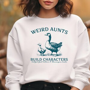 May include: White sweatshirt with the text "WEIRD AUNTS BUILD CHARACTERS" in teal. The graphic features a goose and a gosling. The sweatshirt also has the text "Certified Great Listener & Shenanigans Leader".