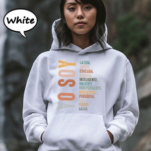 K&ouml;nnte beinhalten: Ein wei&szlig;er Hoodie mit dem Text "YO SOY" in Orange und Gelb und einer Liste spanischer W&ouml;rter in verschiedenen Farben, darunter "LATINA", "MUJER", "EDUCADA", "MAGICA", "INTELIGENTE", "VALIENTE", "INDEPENDIENTE", "ORGULLOSA", "PODEROSA", "LEAL", "FUERTE" und "AMOR".