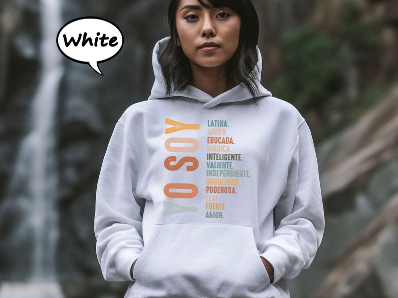 K&ouml;nnte beinhalten: Ein wei&szlig;er Hoodie mit dem Text "YO SOY" in Orange und Gelb und einer Liste spanischer W&ouml;rter in verschiedenen Farben, darunter "LATINA", "MUJER", "EDUCADA", "MAGICA", "INTELIGENTE", "VALIENTE", "INDEPENDIENTE", "ORGULLOSA", "PODEROSA", "LEAL", "FUERTE" und "AMOR".