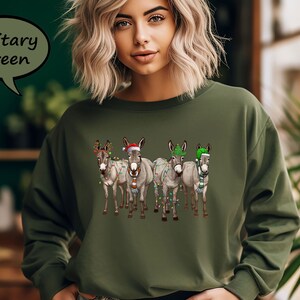 May include: Military green sweatshirt with a graphic of four donkeys wearing Christmas decorations. The donkeys are grey and white, with festive lights, hats, and scarves. The text "Military Green" is in a speech bubble.