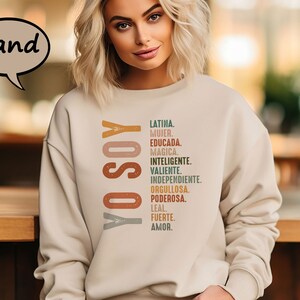 K&ouml;nnte beinhalten: Ein beiger Pullover mit dem Text "YO SOY" in braunen, orangen und grauen Buchstaben. Der Text wird von einer Liste spanischer W&ouml;rter gefolgt, die die Eigenschaften einer Frau beschreiben, darunter "Latina", "Mujer", "Educada", "Magica", "Inteligente", "Valiente", "Independiente", "Orgullosa", "Poderosa", "Leal", "Fuerte" und "Amor."