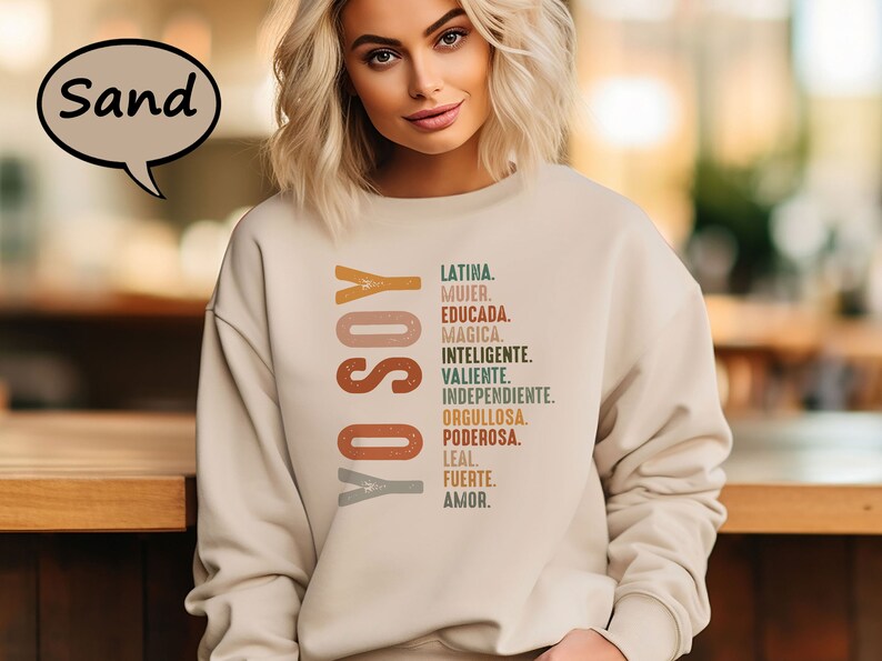 K&ouml;nnte beinhalten: Ein beiger Pullover mit dem Text "YO SOY" in braunen, orangen und grauen Buchstaben. Der Text wird von einer Liste spanischer W&ouml;rter gefolgt, die die Eigenschaften einer Frau beschreiben, darunter "Latina", "Mujer", "Educada", "Magica", "Inteligente", "Valiente", "Independiente", "Orgullosa", "Poderosa", "Leal", "Fuerte" und "Amor."