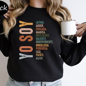 K&ouml;nnte beinhalten: Schwarzes Sweatshirt mit dem Text "YO SOY" in einer bunten Retro-Schrift und einer Liste spanischer W&ouml;rter, die die Eigenschaften einer Frau beschreiben.
