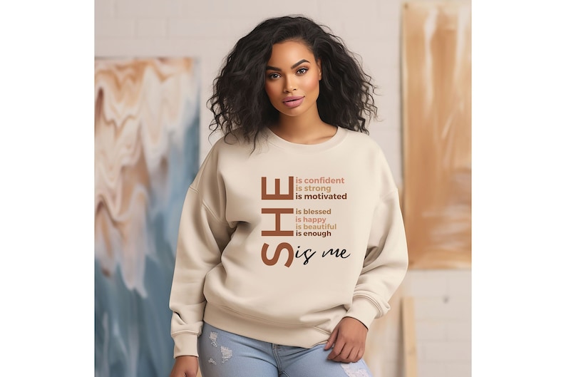 Op de afbeelding: Een beige sweatshirt met de tekst "SHE is confident is strong is motivated is blessed is happy is beautiful is enough is me" gedrukt op de voorkant.