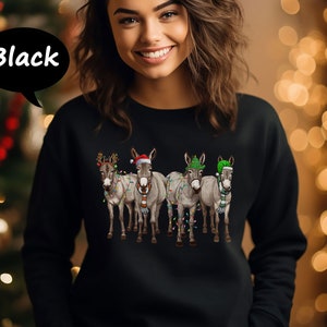 May include: Black sweatshirt with a Christmas-themed design of four donkeys. Each donkey is decorated with festive hats, scarves, and fairy lights. The word "Black" is in a speech bubble.