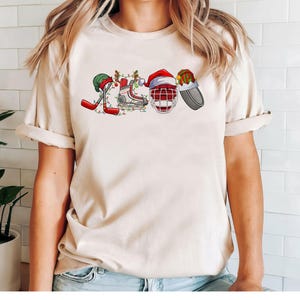 May include: A cream-colored t-shirt with a Christmas-themed hockey design. The design features a hockey stick, skates, a hockey helmet, and a stocking cap.
