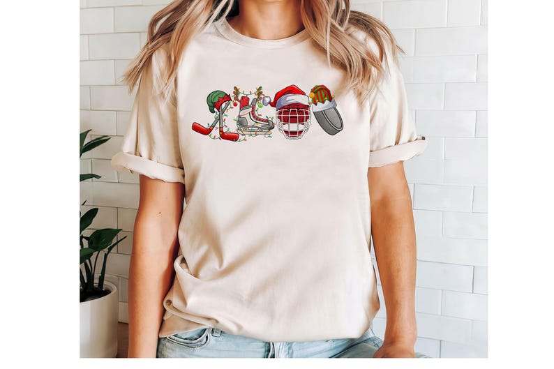 May include: A cream-colored t-shirt with a Christmas-themed hockey design. The design features a hockey stick, skates, a hockey helmet, and a stocking cap.