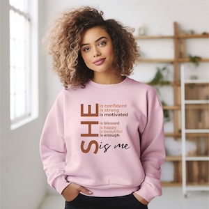 Op de afbeelding: Een roze sweatshirt met de tekst "SHE is confident is strong is motivated is blessed is happy is beautiful is enough is me" in bruin gedrukt op de voorkant.