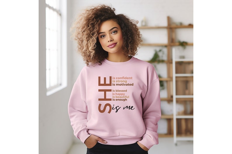 Op de afbeelding: Een roze sweatshirt met de tekst "SHE is confident is strong is motivated is blessed is happy is beautiful is enough is me" in bruin gedrukt op de voorkant.