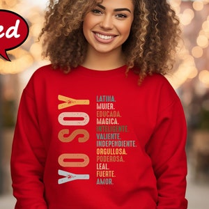 K&ouml;nnte beinhalten: Roter Pullover mit dem Text "YO SOY" in einer vertikalen, bunten Retro-Schrift. Dem Text folgt eine Liste spanischer W&ouml;rter, die die Eigenschaften einer Frau beschreiben, darunter Latina, Mujer, Educada, Magica, Inteligente, Valiente, Independiente, Orgullosa, Poderosa, Leal, Fuerte, Amor.