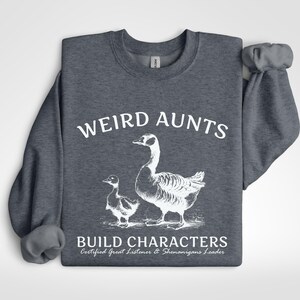 May include: Dark gray sweatshirt with the text "WEIRD AUNTS" above a graphic of a goose and gosling. Below the image, the text reads "BUILD CHARACTERS" and "Certified Great Listener & Shenanigans Leader."